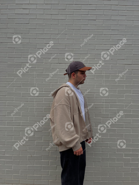 a young man in simple clothes stands sideways against the background of a brick wall